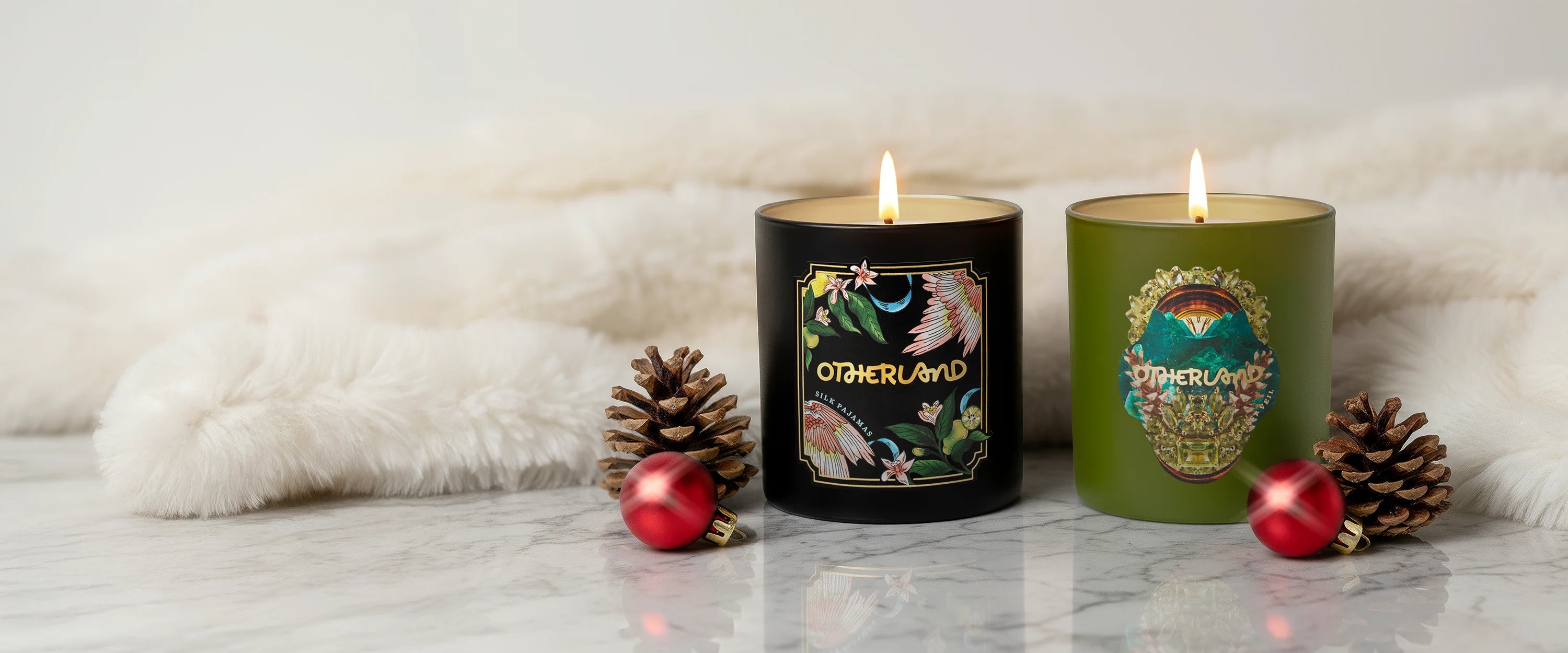 Two lit Otherland candles on a white surface with decorative pinecones and berries.