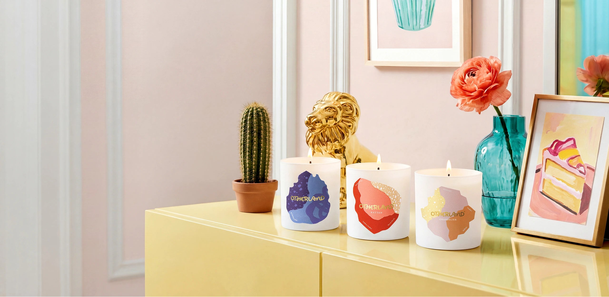 Decorative candles with colorful designs on a yellow surface, surrounded by small plants and framed artwork.