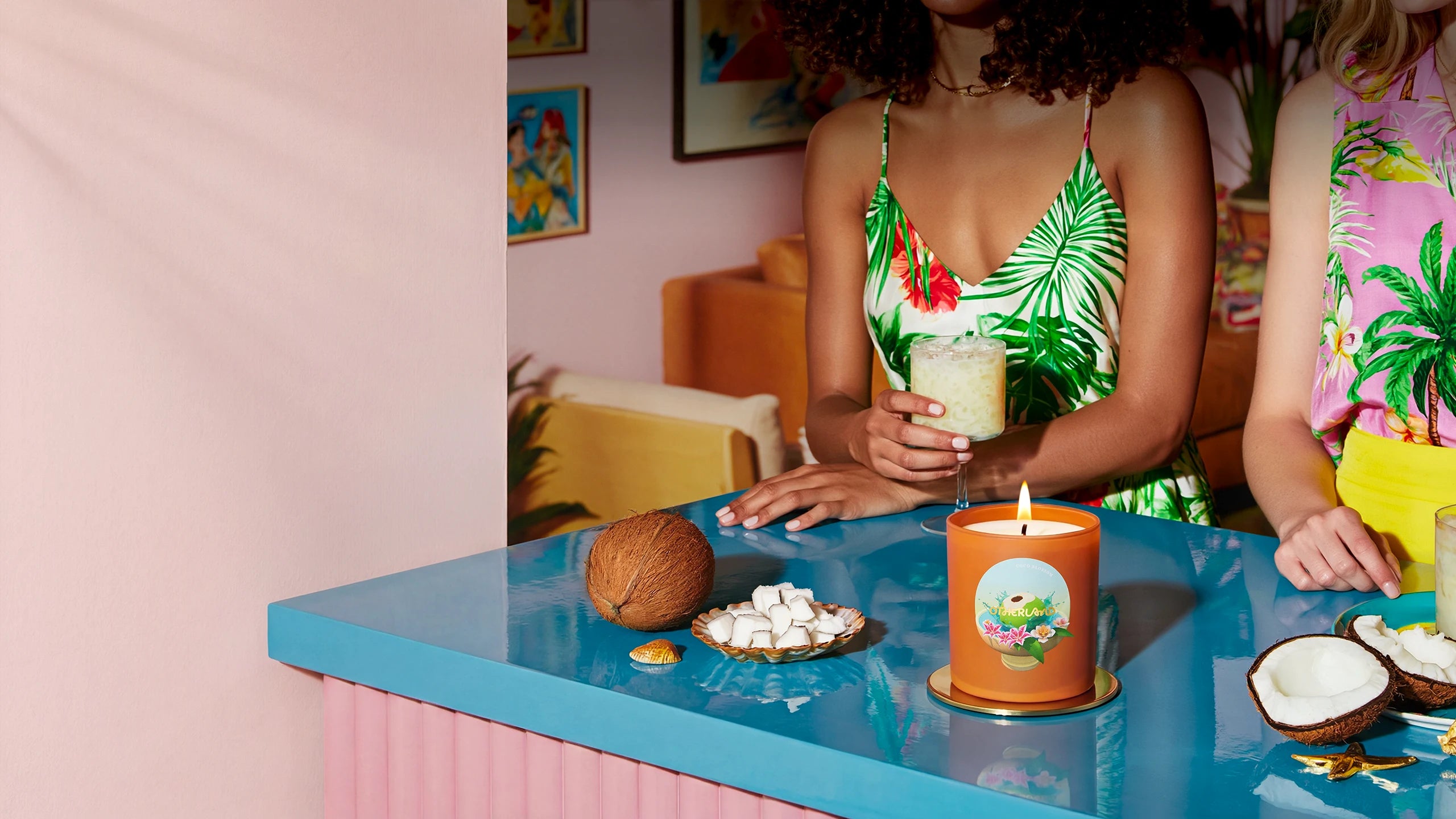 Two women in tropical-themed outfits standing behind a counter with a candle and snacks.