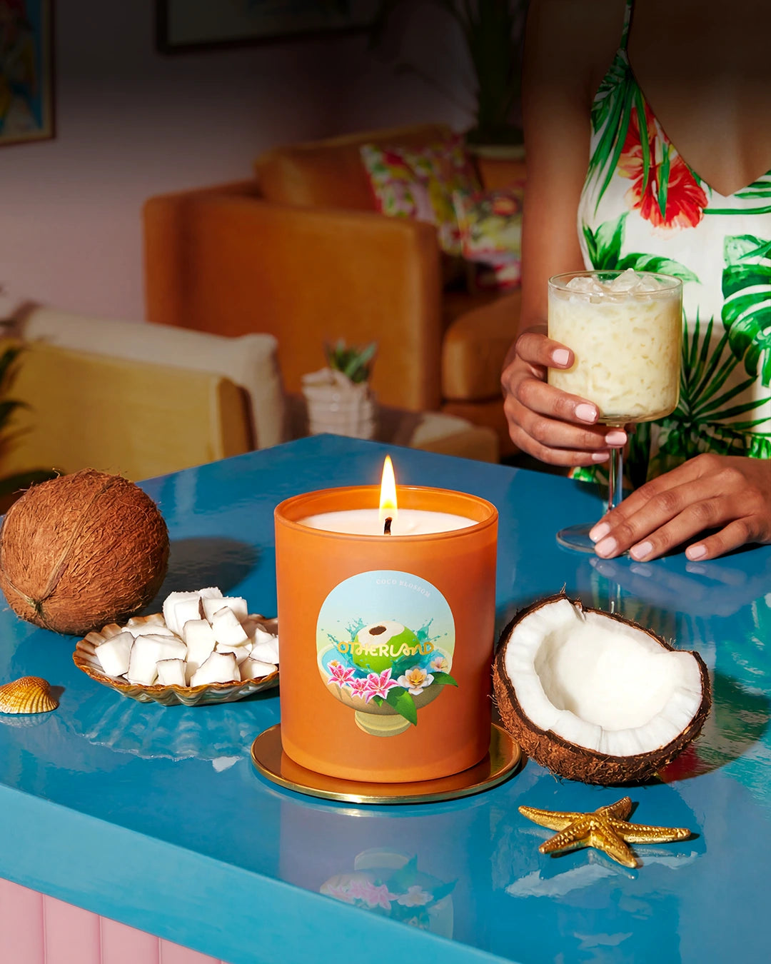 Orange candle with a label on a blue table, surrounded by coconuts and a glass of a drink.