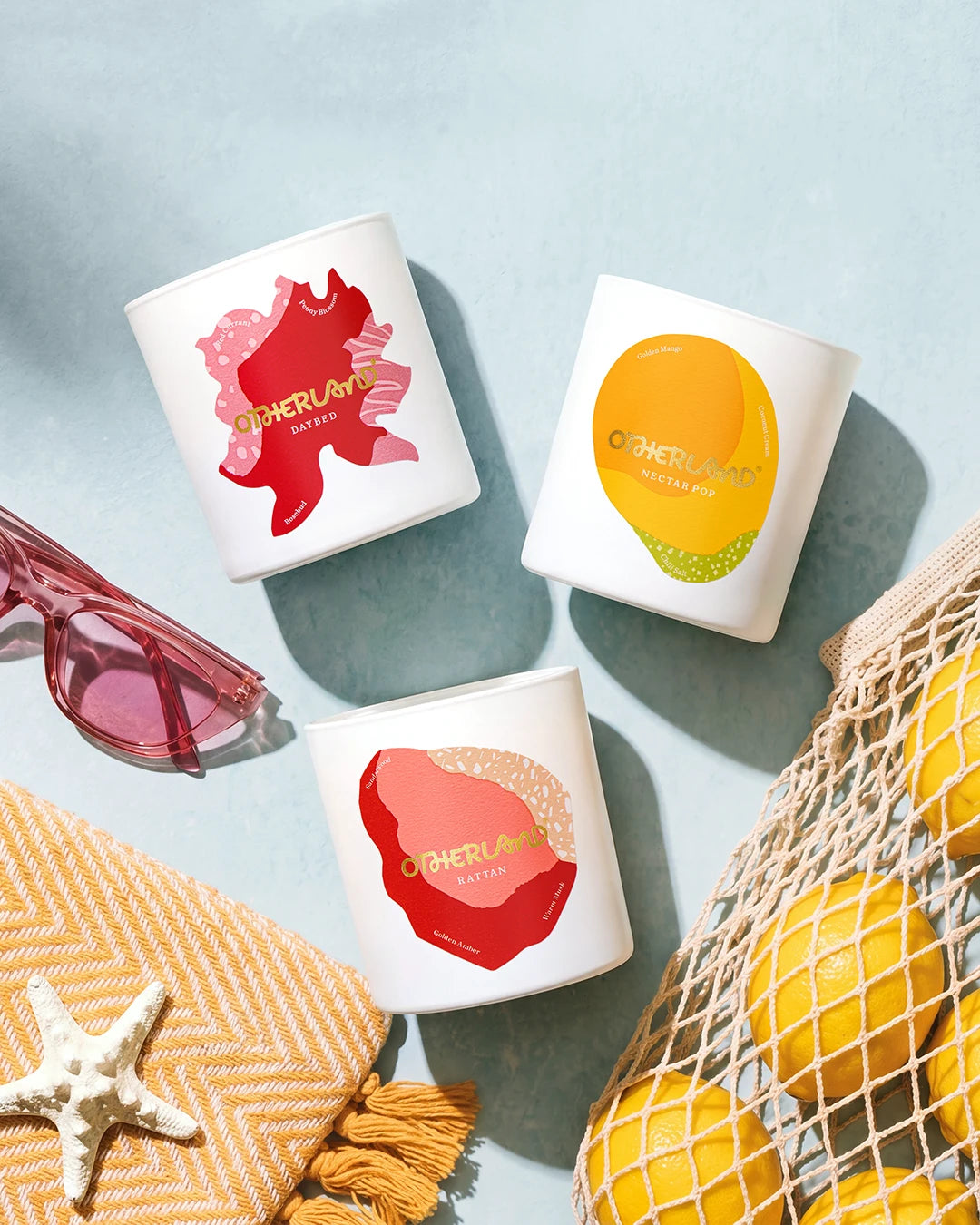 Three candles with colorful designs on a light blue surface, surrounded by sunglasses, a yellow bag, and a starfish.
