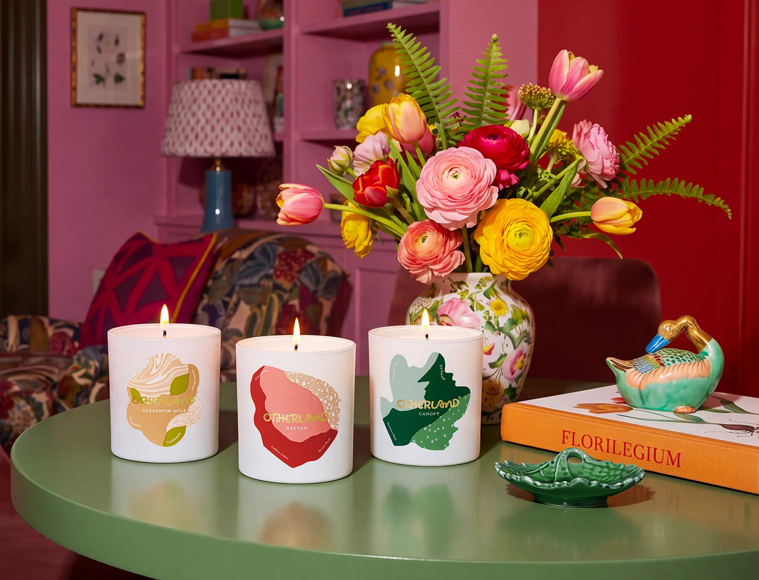 Three candles with decorative designs on a table, accompanied by a vase of flowers and a book titled 'Florilegium'.