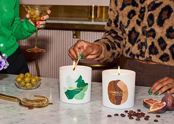 Two candles with decorative labels on a table with figs and olives.