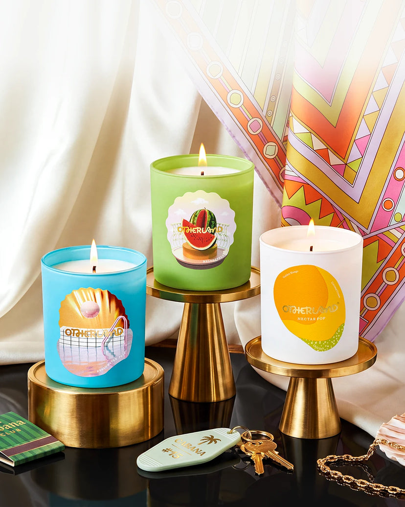 Three lit candles in colorful cylindrical jars displayed on gold pedestal stands against draped white fabric and a patterned textile backdrop. The candles have illustrated fruit and citrus labels, with one blue jar, one green jar, and one white jar.
