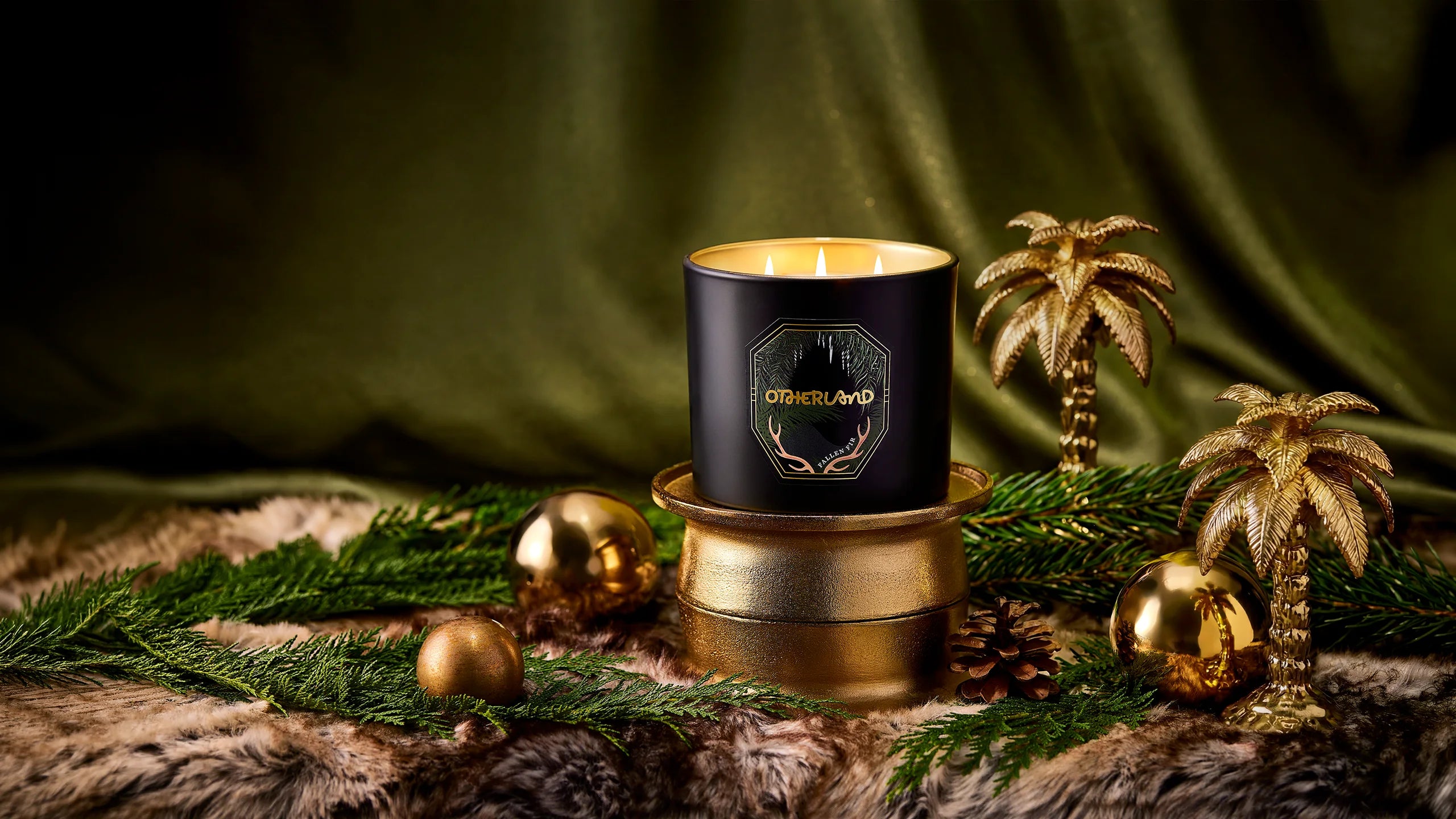 A black jar candle with gold lettering sits on a gold-toned base, styled with green pine branches, gold ornaments, and small palm tree decorations against a dark green draped fabric background.