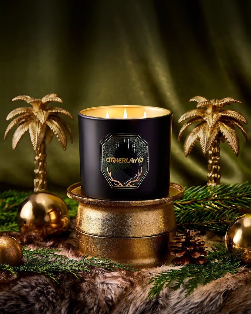 A black scented candle in a round jar with gold lettering reading 'OTHERLAND' and antler graphics, displayed on a gold-toned pedestal. The candle is styled with miniature gold palm trees, green pine branches, pinecones, and gold ornaments against a dark green draped background.