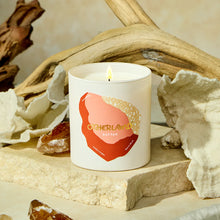 Candle with 'Otherland' branding on a natural setting with driftwood and shells.