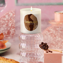 Candle with Otherland on a decorative surface with coffee beans and pastries.