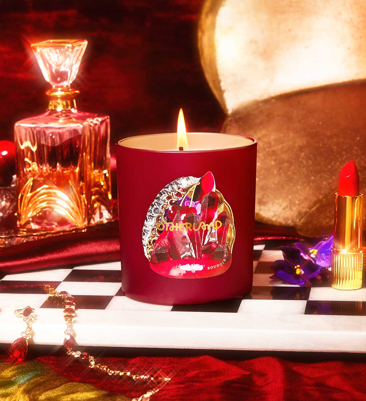 Red candle with a decorative label on a checkered surface, surrounded by a bottle, lipstick, and other items.