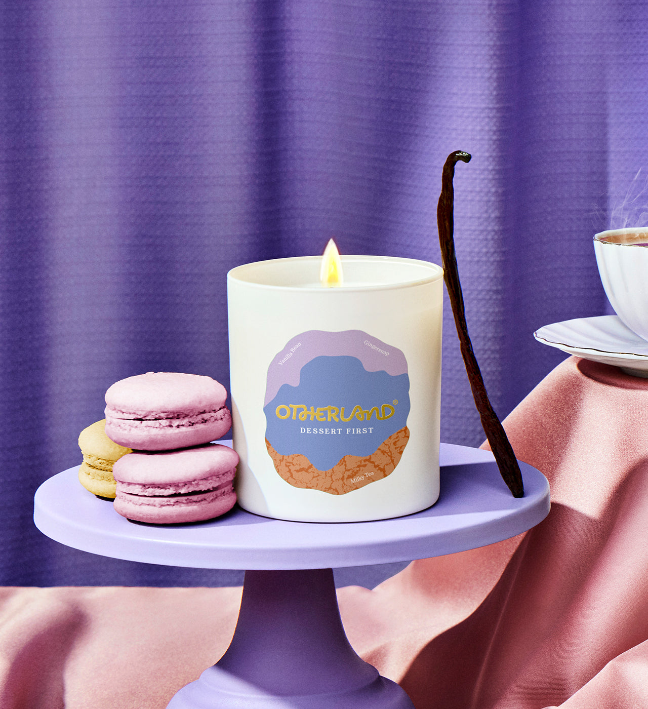 Candle with a dessert design on a purple stand with macarons and a vanilla bean against a purple curtain. - View 2
