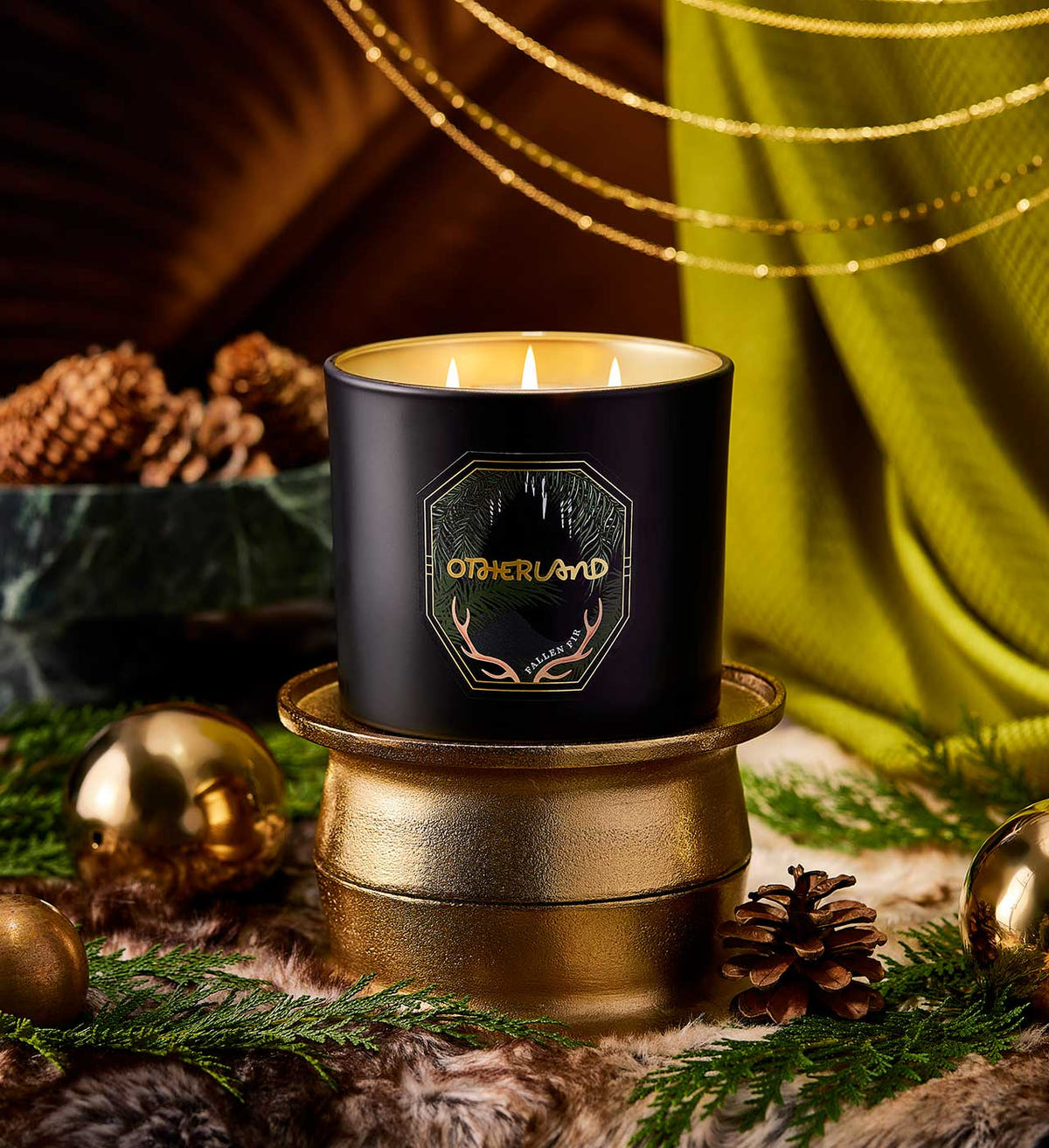 Black candle with 'Otherland' branding on a decorative surface with greenery and gold accents.