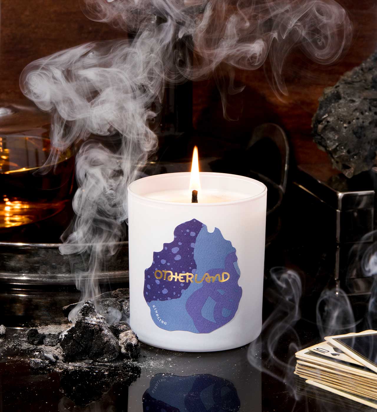 Candle with 'Otherland' logo on a dark background with smoke and rocks.